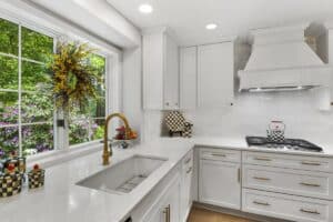 Bright, modern kitchen remodel with white cabinets, gold hardware, and a farmhouse sink under a large window adorned with a floral wreath. Decorative checkered containers and a gas stove sit on the white countertop.