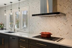 Modern kitchen remodel with black cabinets, white countertop, herringbone tile backsplash, stainless steel range hood, gas cooktop with a red pot, under-mount sink, and three pendant lights by a window.