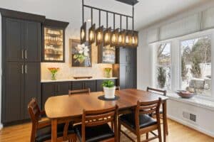 A modern dining room, refreshed by a recent kitchen remodel, features a wooden table, six chairs, potted plant centerpiece, dark cabinets, wall art, pendant lighting, and a large window overlooking the snowy yard.