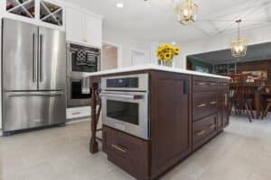 Modern kitchen remodel with stainless steel appliances, a large island featuring a built-in oven and dark wood cabinets, white countertops, sunflowers in a vase, and pendant lights hanging above.