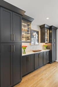 A modern kitchen remodel featuring matte black cabinets, glass display shelves with glassware, a light marble countertop, wood flooring, two vases of red tulips, and a framed flower artwork on the wall.