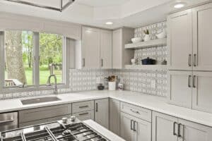 Modern kitchen remodel with light gray cabinets, white countertops, patterned white tile backsplash, open shelves displaying dishes, a stove in the foreground, and a large window overlooking a green yard with trees.