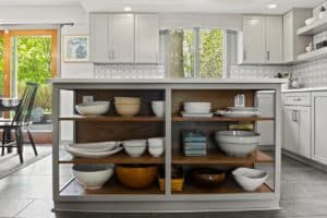 Open kitchen shelves under an island display neatly arranged bowls, plates, and dishes. This kitchen remodel features white cabinetry, a window with a tree view, and a sliding glass door for a bright, modern atmosphere.