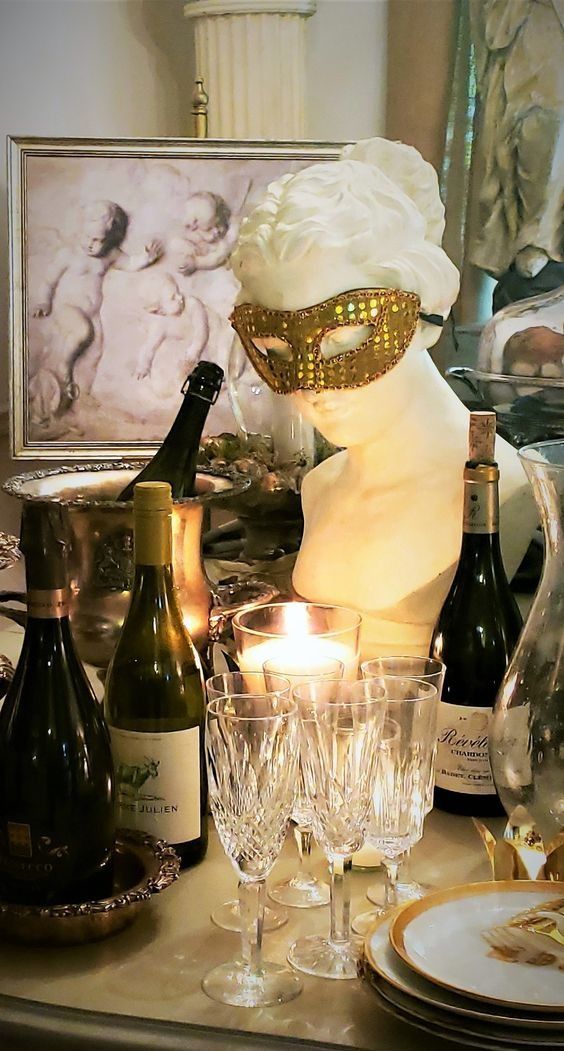 A white bust of a woman with a gold mask and a lit candle surrounded by wine glasses and bottles.