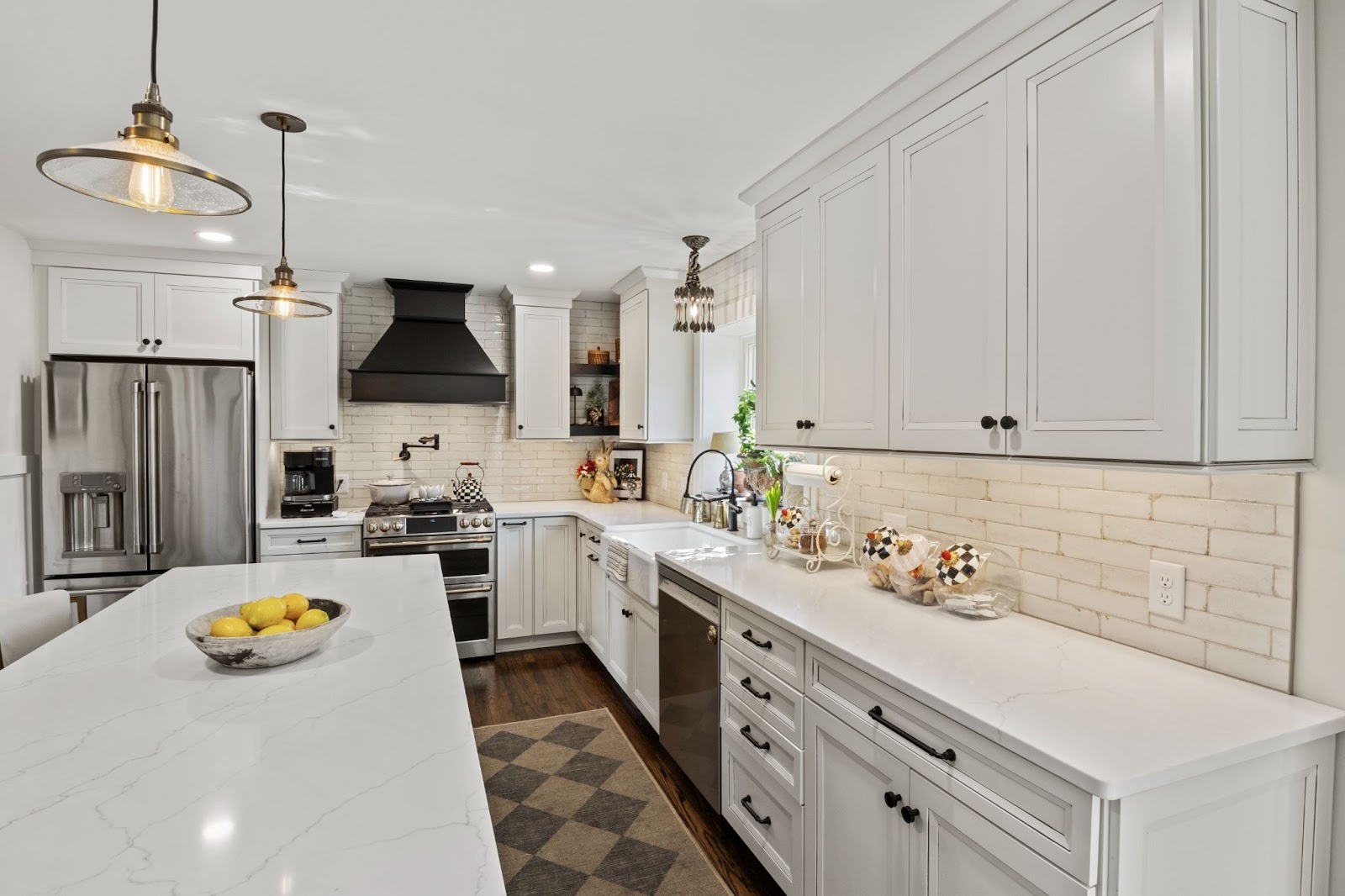 A modern white kitchen with a large island, white cabinets, and stainless steel appliances. The kitchen features a bowl of fruit on the island.