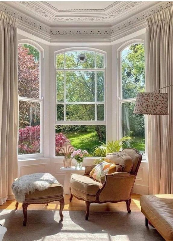 A comfortable living room with a bay window overlooking a garden. The room is furnished with a chair, ottoman, and table.