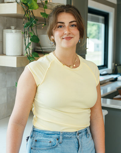A young woman wearing a yellow top and jeans standing in a kitchen. She is smiling and looking at the camera.