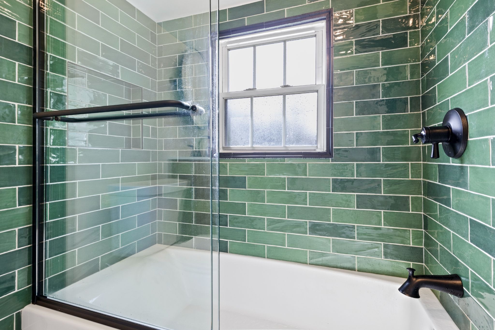 A bathroom shower with green tile walls and a window. The shower has a white tub and black fixtures.
