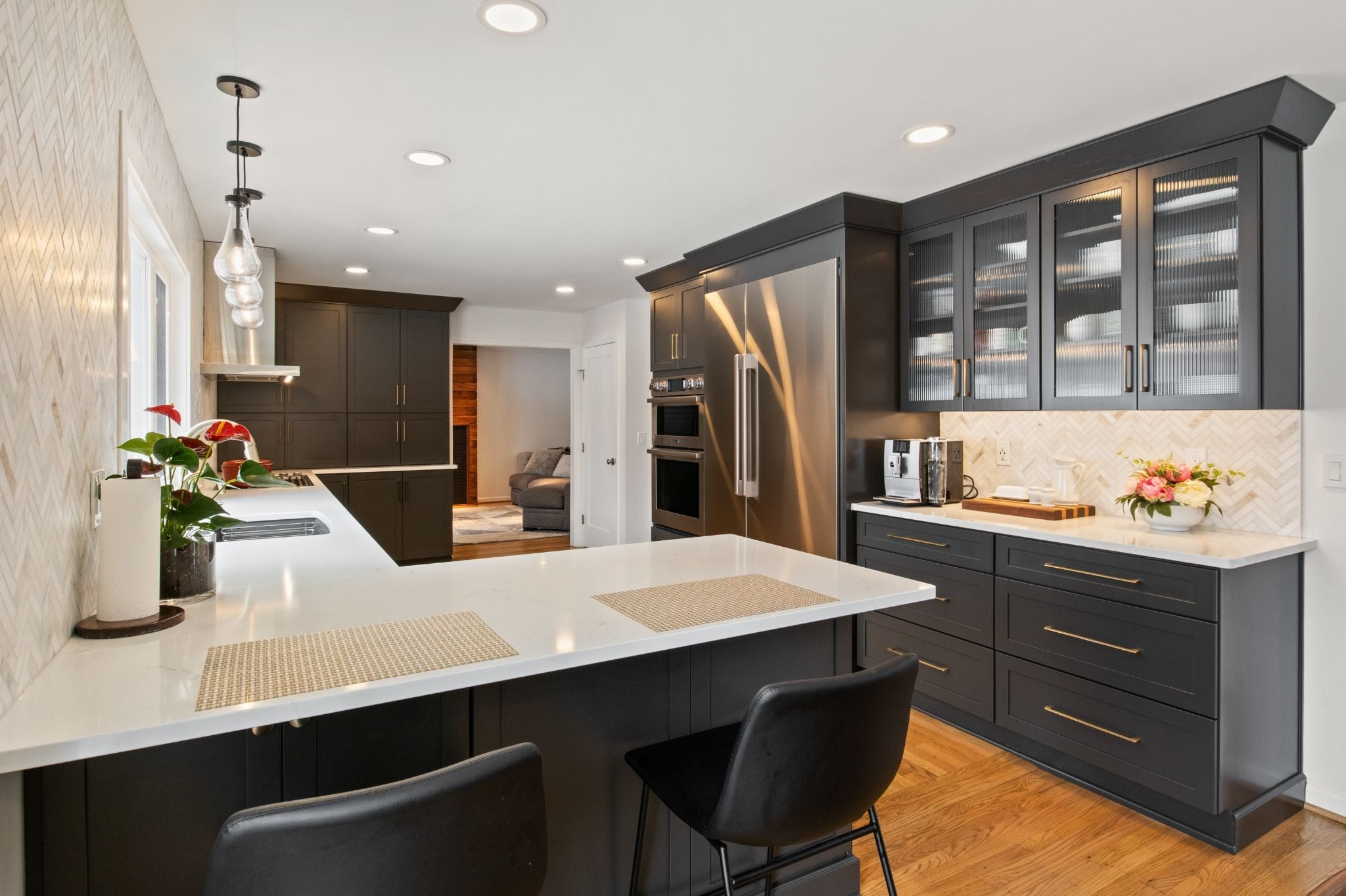 A modern kitchen with dark cabinets, white countertops, and a large island. The kitchen features a stainless steel refrigerator and a sink.