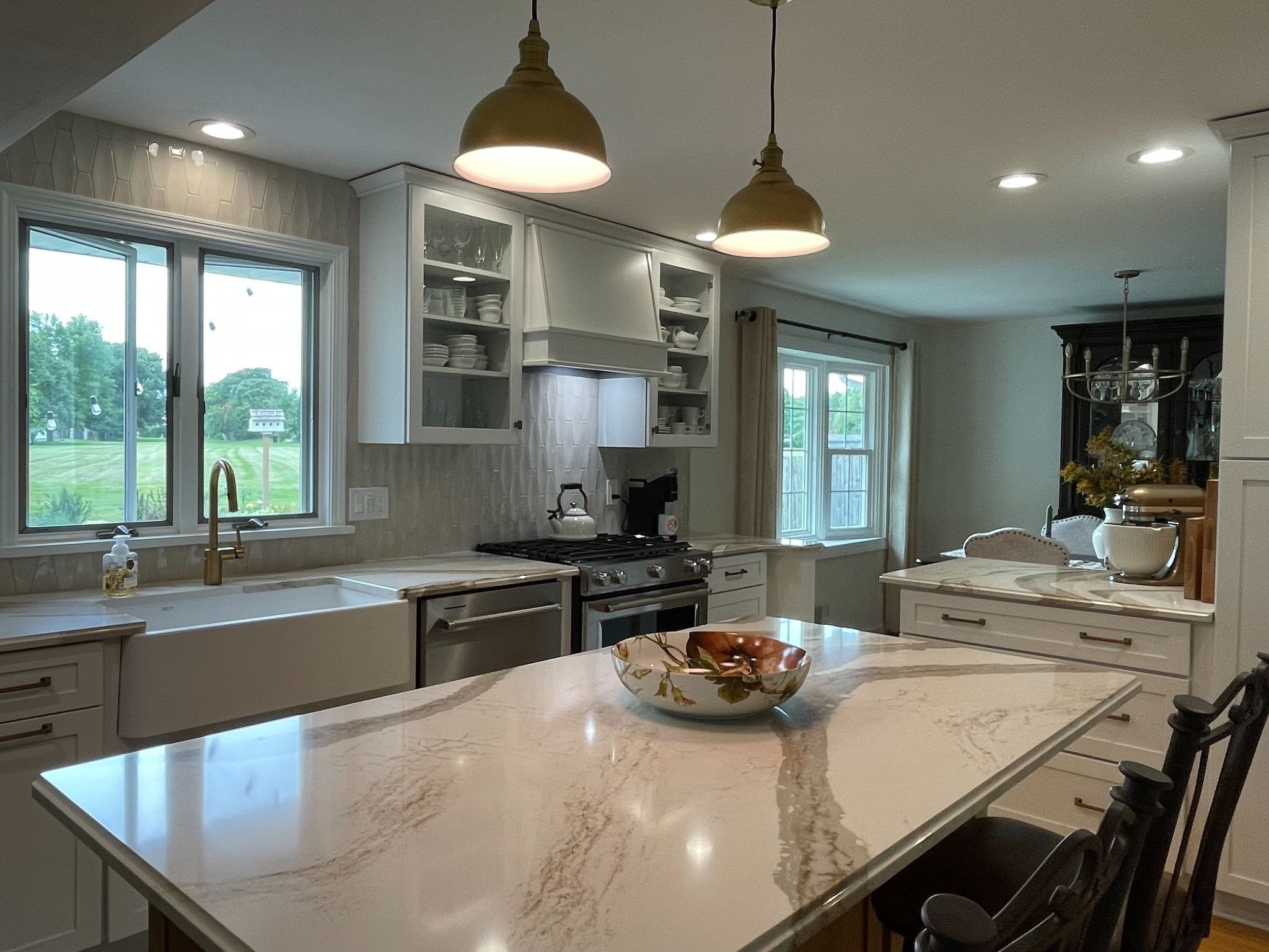 A modern kitchen with a large island featuring a marble countertop and a bowl of fruit. The kitchen is well-lit with pendant lights.