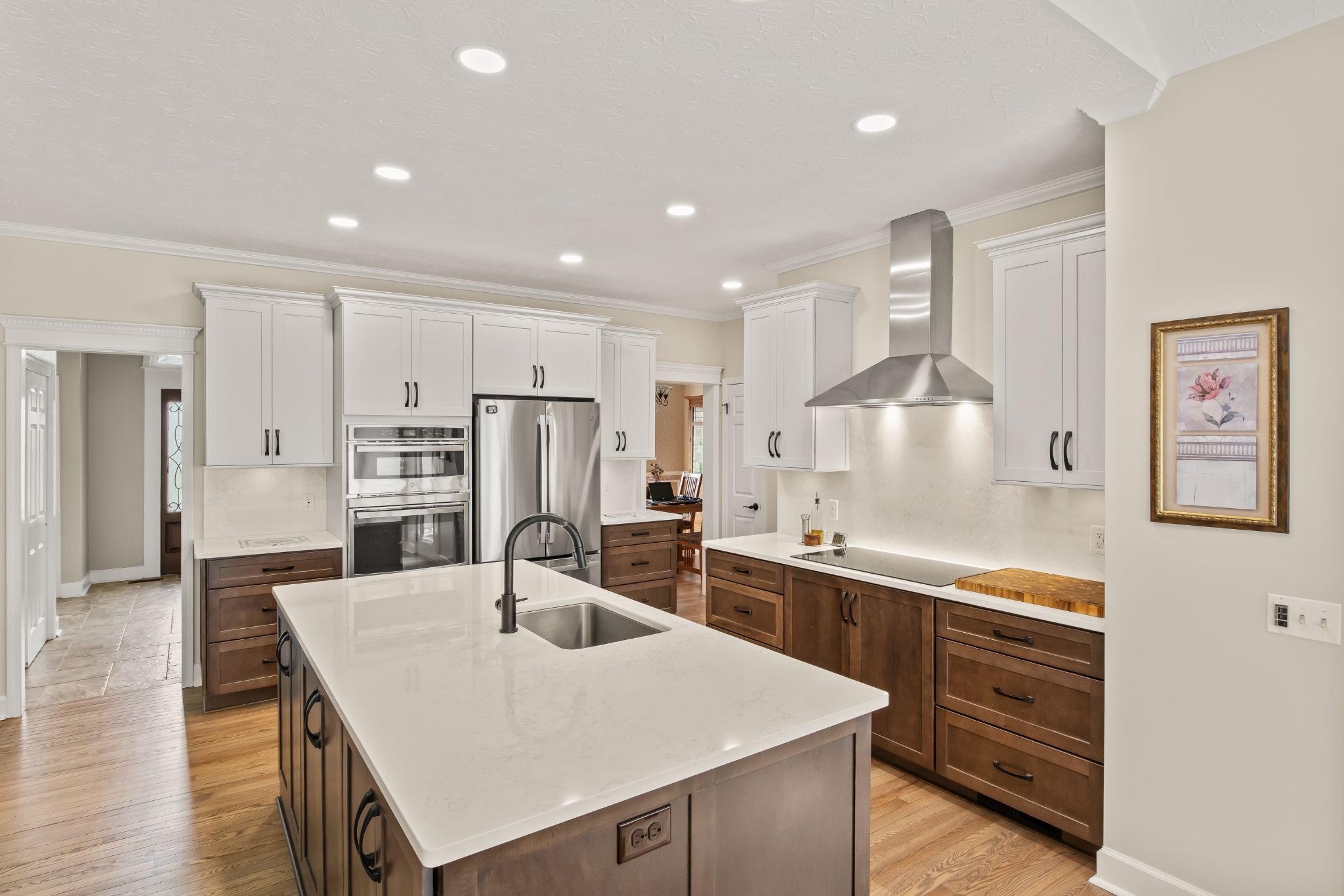 A modern kitchen with a large island, white countertops, and stainless steel appliances. The kitchen features dark wood cabinets and a stainless steel range hood.