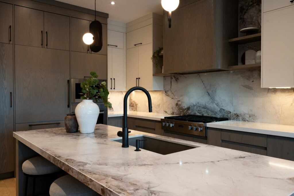 A modern kitchen with a large marble countertop, dark gray cabinets, and a black faucet. The kitchen is well-lit with pendant lights.