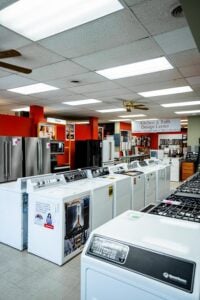 Rows of Maytag washing machines on display in a home appliance store with bright ceiling lighting.