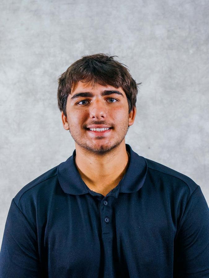A smiling young man with brown hair wearing a navy blue polo shirt against a gray background. The image appears to be a professional headshot or profile photo.