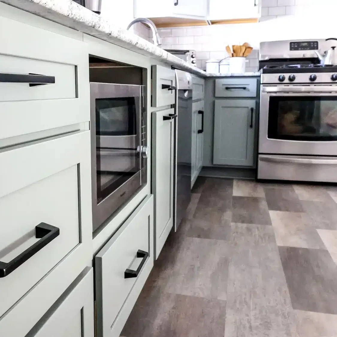Modern kitchen remodel with light green cabinets, black handles, a built-in microwave, gas stove with oven, and mixed light and dark gray flooring. White countertops display kitchen items in the background for a fresh update.