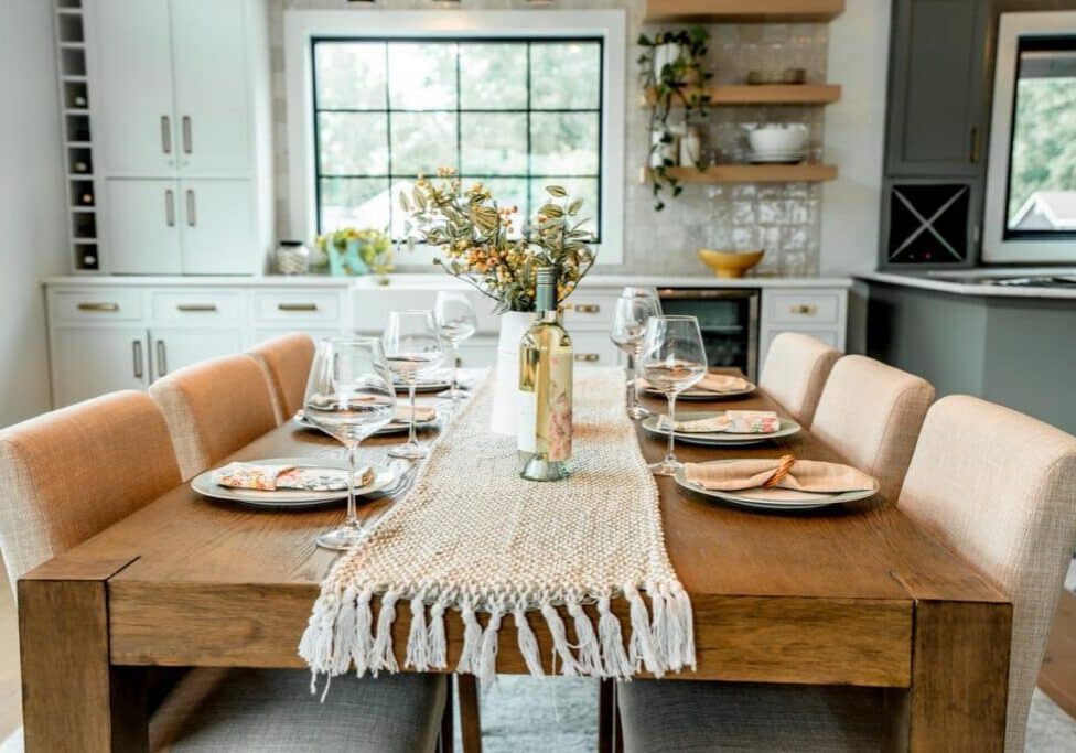 A wooden dining table set for six with wine glasses, plates, and a floral centerpiece creates a welcoming scene in a bright, modern kitchen—just like the elegant designs showcased in the Webster Showroom.