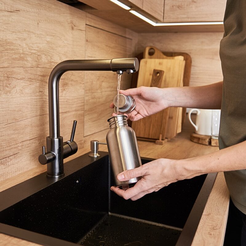 A person fills a stainless steel water bottle with water from one of the best kitchen faucets over a black sink, with wooden cabinets and cutting boards in the background.