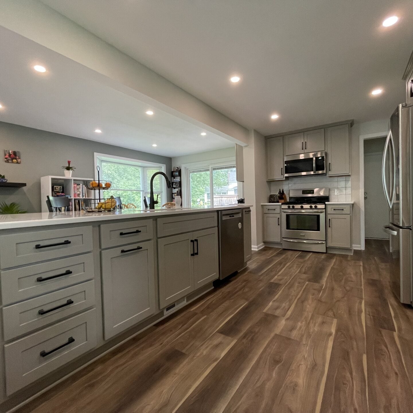 Modern kitchen remodel with gray cabinets, stainless steel appliances, an island with a sink and dishwasher, wood-look flooring, recessed lighting, and large windows providing natural light.
