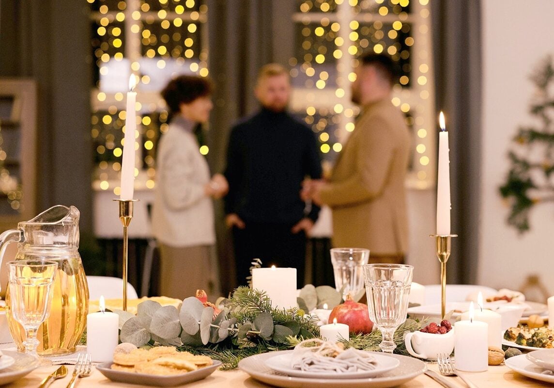 A festively decorated dinner table with candles, food, and glasses in the foreground sets the scene for stress-free entertaining, while three people stand and talk in the softly lit background with string lights in the windows.