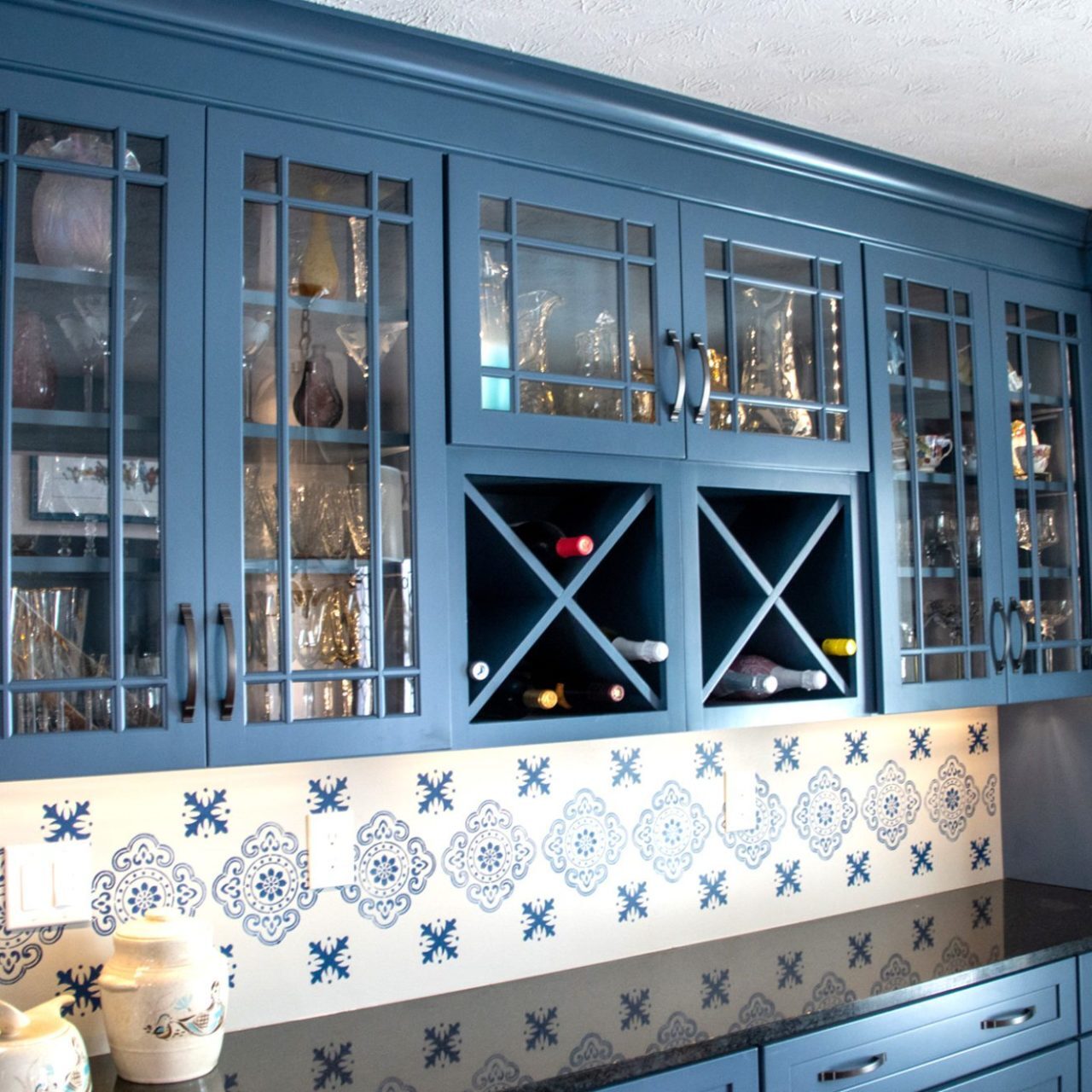 Blue kitchen cabinets with glass doors display dishes and glasses, perfect for your kitchen remodel. A backsplash features blue and white decorative tiles. Built-in wine racks hold bottles, while under-cabinet lighting highlights decorative pitchers on the countertop. Blue kitchen cabinets with glass doors display dishes and glasses, perfect for your kitchen remodel. A backsplash features blue and white decorative tiles. Built-in wine racks hold bottles, while under-cabinet lighting highlights decorative pitchers on the countertop.
