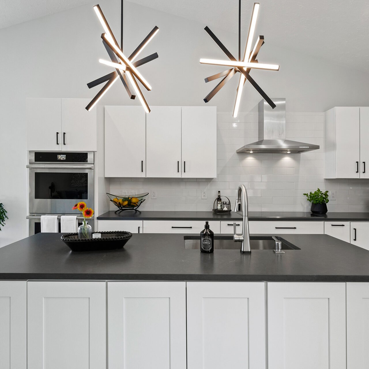Modern kitchen remodel with white cabinets, black countertops, stainless steel appliances, and two geometric pendant lights above a large island with a sink. A window lets in natural light, and plants decorate the space. Modern kitchen remodel with white cabinets, black countertops, stainless steel appliances, and two geometric pendant lights above a large island with a sink. A window lets in natural light, and plants decorate the space.