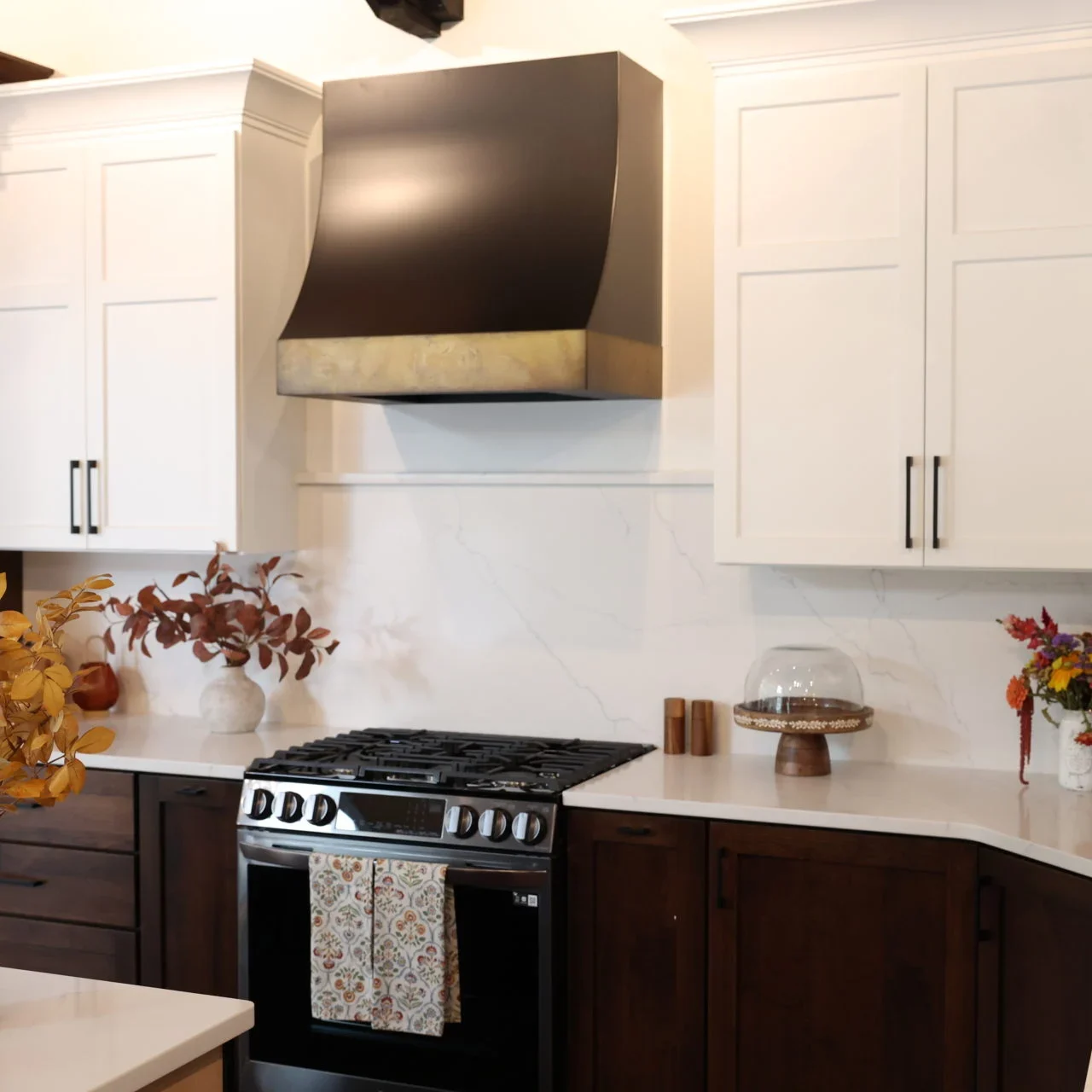 A modern kitchen remodel with white upper cabinets, dark lower cabinets, a black and brass range hood over the stove, and fall-themed floral arrangements on the countertops. A dish towel hangs on the oven door. A modern kitchen remodel with white upper cabinets, dark lower cabinets, a black and brass range hood over the stove, and fall-themed floral arrangements on the countertops. A dish towel hangs on the oven door.