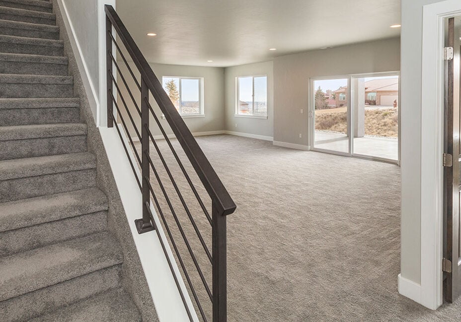 A spacious, carpeted basement with neutral walls, large windows letting in natural light, a wooden door, and a staircase with a black metal railing leading upstairs—perfect for relaxing after a kitchen remodel.