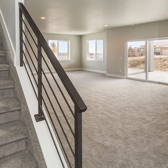 A spacious, carpeted basement with neutral walls, large windows letting in natural light, a wooden door, and a staircase with a black metal railing leading upstairs—perfect for relaxing after a kitchen remodel. A spacious, carpeted basement with neutral walls, large windows letting in natural light, a wooden door, and a staircase with a black metal railing leading upstairs—perfect for relaxing after a kitchen remodel.
