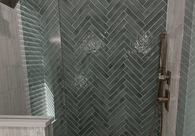 A modern shower with glossy dark green herringbone tile, a rainfall showerhead on the left, and a vertical bar with a handheld shower and controls on the right—perfect inspiration for your next bath or kitchen remodel. A glass enclosure corner is visible.