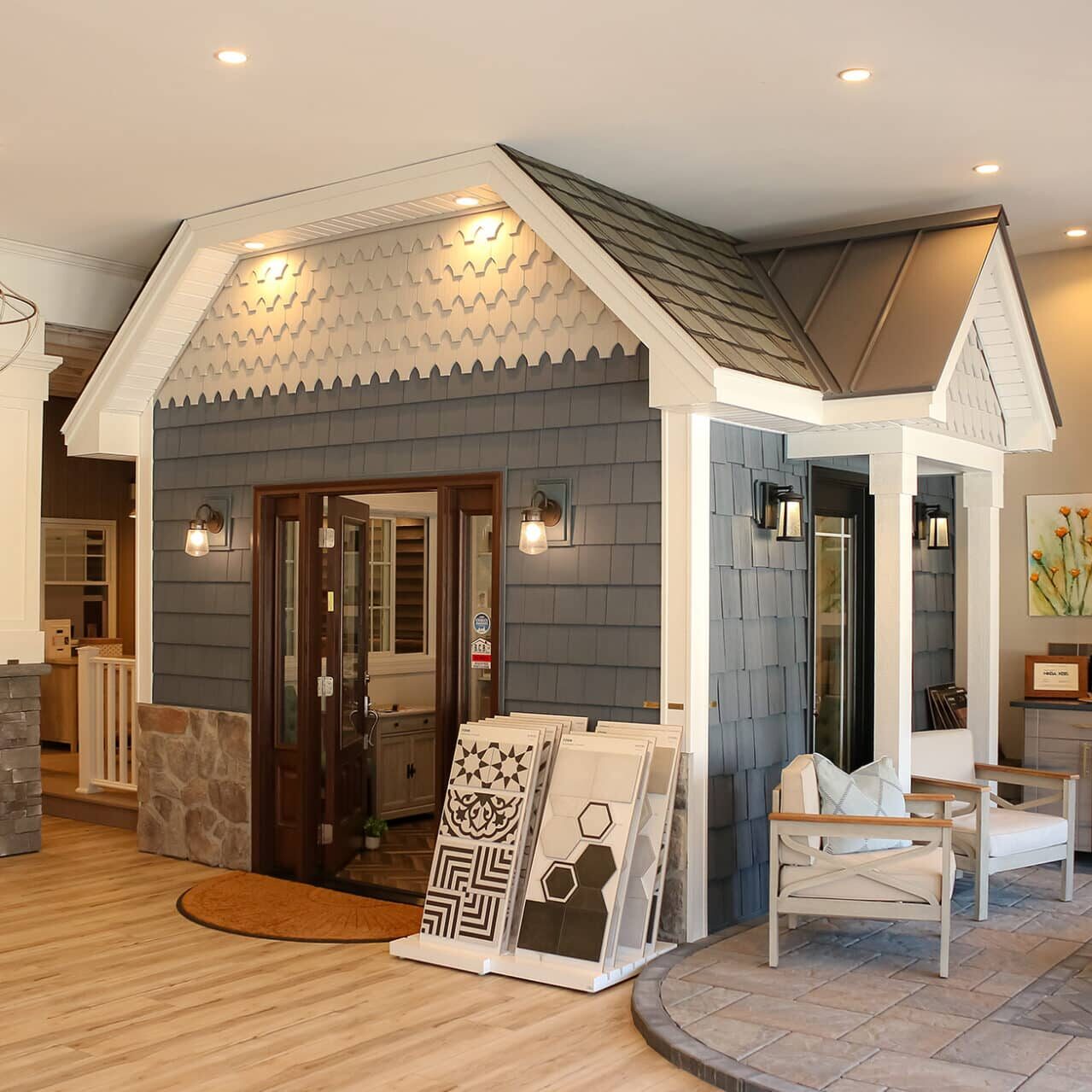 A showroom with a small house display featuring gray siding, a shingled roof, and double doors. Inside, see fireplace, chairs, flooring samples, decorative lighting, and inspiration for your next kitchen remodel in the spacious, well-lit interior.