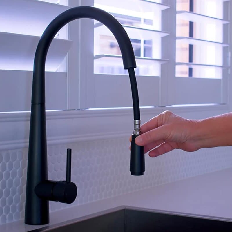 A hand pulls down the spray nozzle of a modern black kitchen faucet—one of the best types of kitchen faucets—over a sink, with white tile backsplash and window shutters in the background.