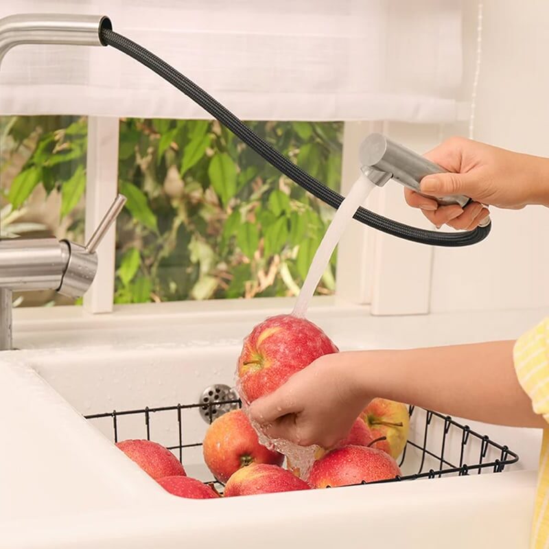 A person in a yellow shirt uses one of the best kitchen faucets—a pull-down model—to wash red apples in a sink, holding one apple under running water, with more apples in a black wire basket below.