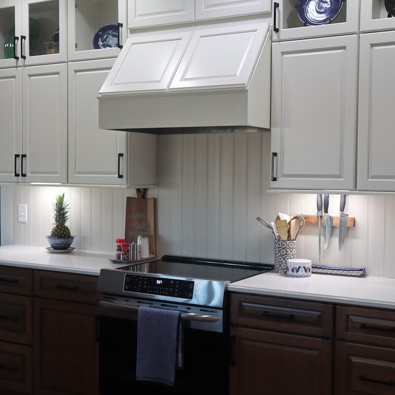 A modern kitchen remodel features white upper cabinets, dark lower cabinets, a stainless steel stove with a towel hanging on the handle, a knife rack, utensils, a small potted plant, and decorative plates on display.