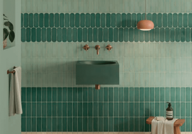 A stylish bathroom reminiscent of a luxe kitchen remodel, with green vertical tiles, a dark green sink, wall-mounted brass faucet, coral wall light, towel on a hook, small wooden stool with accessories, and a leafy plant partially visible on the left.