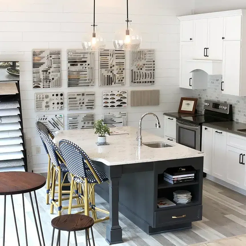 A modern kitchen showroom perfect for your kitchen remodel, featuring white cabinets, a large island with a marble countertop, blue and gold barstools, pendant lights, and display walls showcasing various cabinet handles and samples.