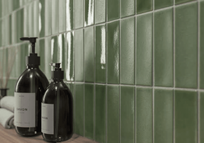 Two dark pump soap bottles sit on a wooden surface in front of a wall of glossy green square tiles—an elegant detail to inspire your next kitchen remodel. A rolled towel and small container are partially visible in the background.