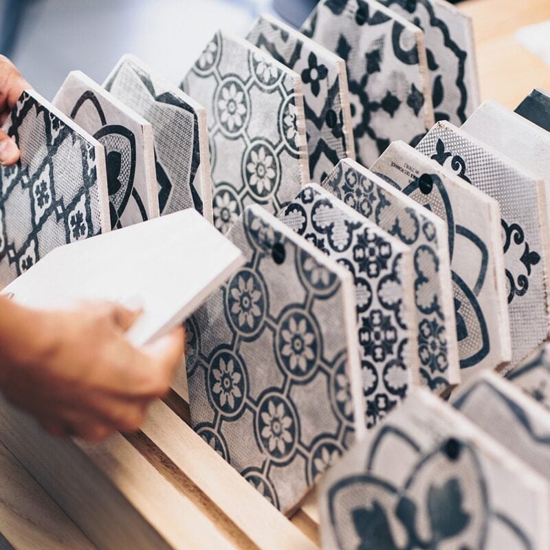 A person selects from a wooden display rack holding various black-and-white patterned ceramic tiles, featuring geometric and floral designs—perfect choices for stylish waterproof flooring.
