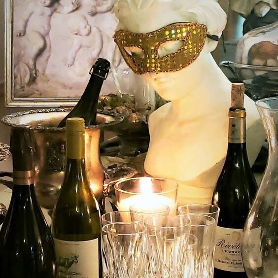 A white bust of a woman with a gold mask and a lit candle surrounded by wine glasses and bottles.