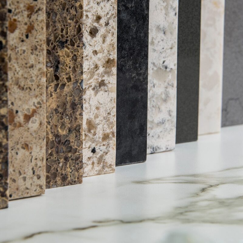 A row of upright countertop samples in various colors and textures, mostly shades of brown, beige, black, and gray, displayed on a white surface with gray veining—ideal for pairing with waterproof flooring options.
