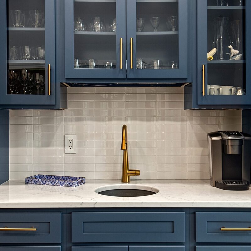 A modern kitchen area with blue cabinets, gold handles, a gold faucet over a small round sink—perfect as a wet bar. Features include a white tile backsplash, marble countertop, coffee maker, and patterned tissue box.