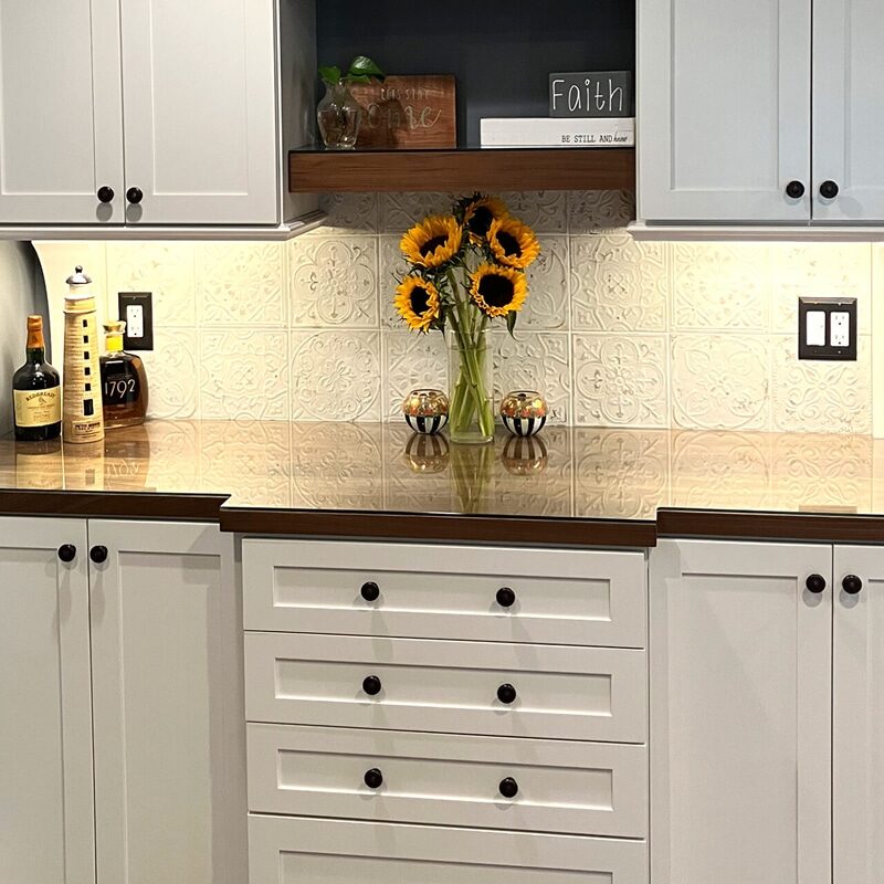 A kitchen counter with white cabinets, a dark wood countertop, a vase of sunflowers, bottles, and decorative items creates a charming home bar. A patterned white backsplash and a small wooden shelf with decor sit above the counter.