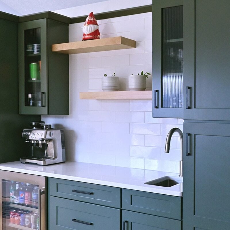 A modern kitchen nook with dark green cabinets, white countertop, built-in mini fridge, espresso machine, and open wooden shelves holding dishes, potted plants, and a decorative Santa—perfect for stress-free entertaining or holiday party prep. White tile backsplash.