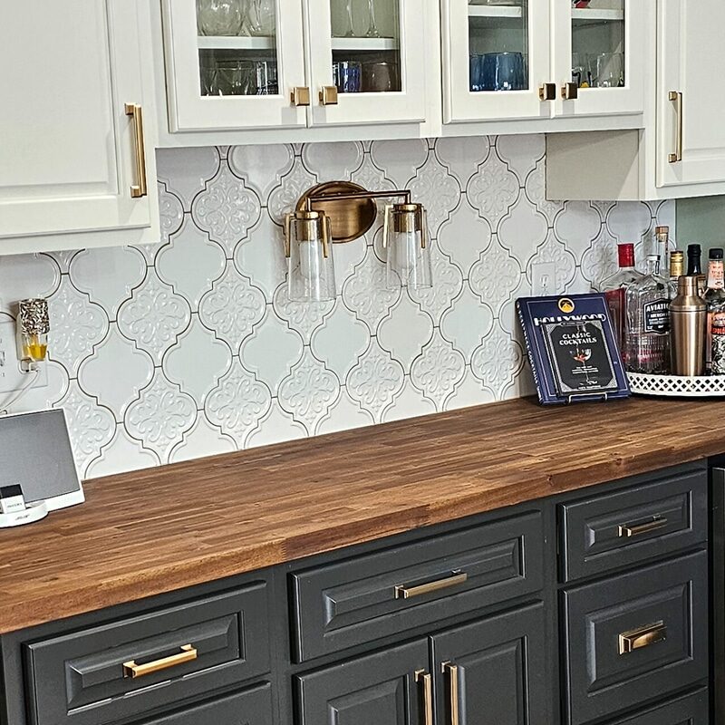 A stylish home bar setup features a wood kitchen counter, dark lower cabinets with gold handles, white glass-front uppers, and a glass-front mini fridge. Bottles, a tray, a cookbook, and a speaker complete this elegant dry bar space.