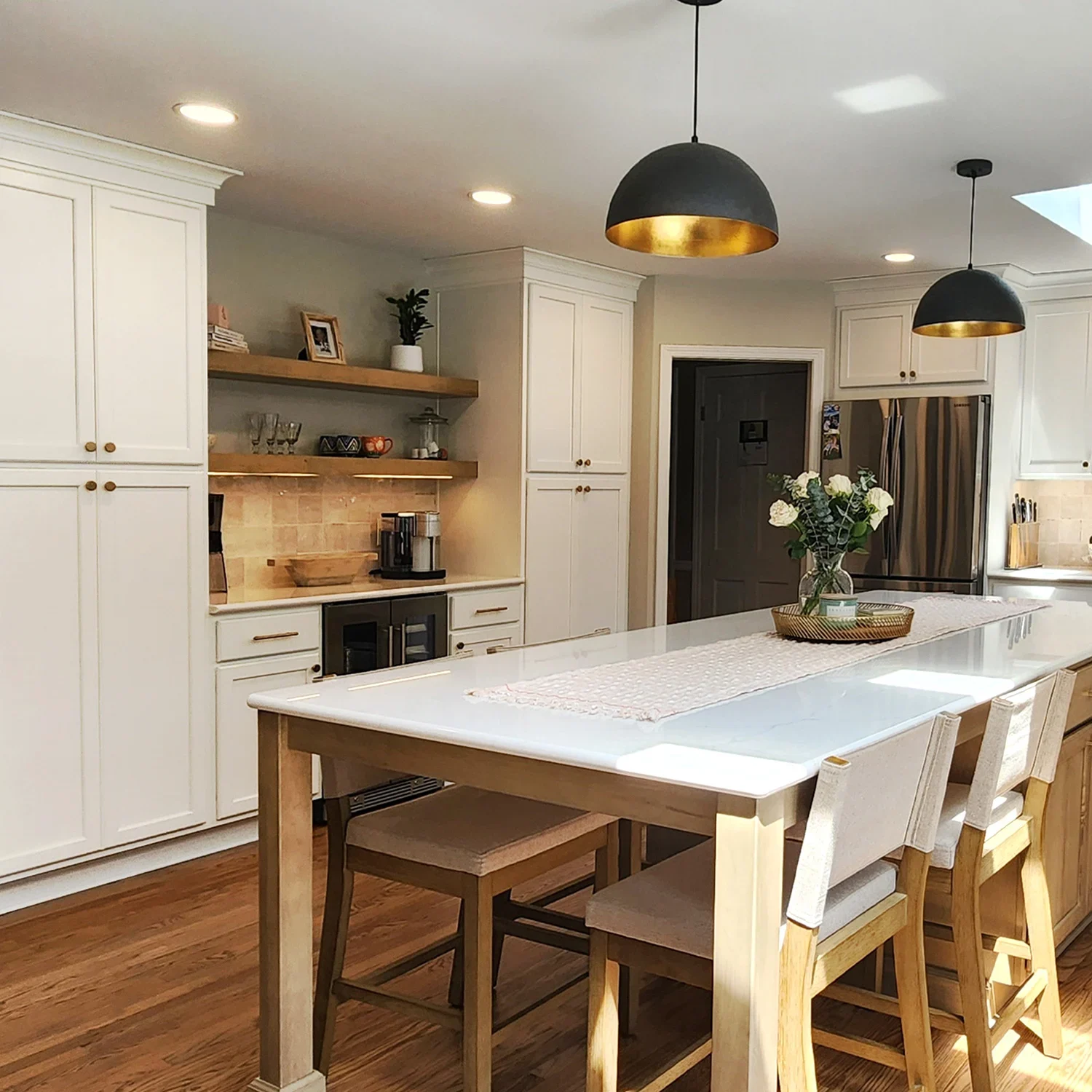 Bright, modern kitchen remodel with white cabinets, light wood flooring, a large island with a white countertop, beige cushioned chairs, black pendant lights, and decorative flowers on the island. Bright, modern kitchen remodel with white cabinets, light wood flooring, a large island with a white countertop, beige cushioned chairs, black pendant lights, and decorative flowers on the island.