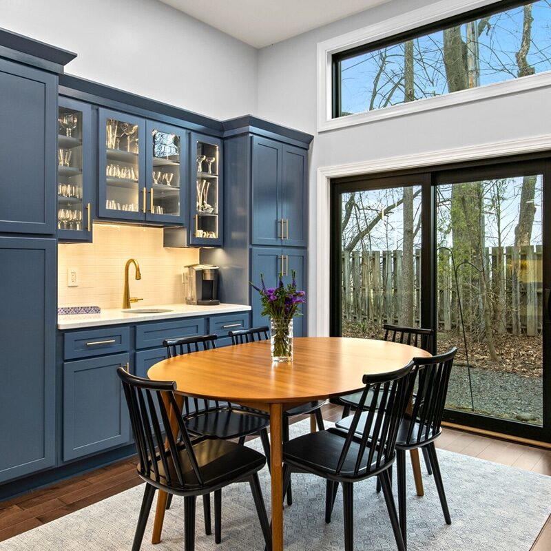 A modern kitchen with blue cabinets, a stylish wet bar, a round wooden table with four black chairs, under-cabinet lighting, and large windows framing trees outside. A vase of flowers sits on the table, with plants on a white stand nearby.