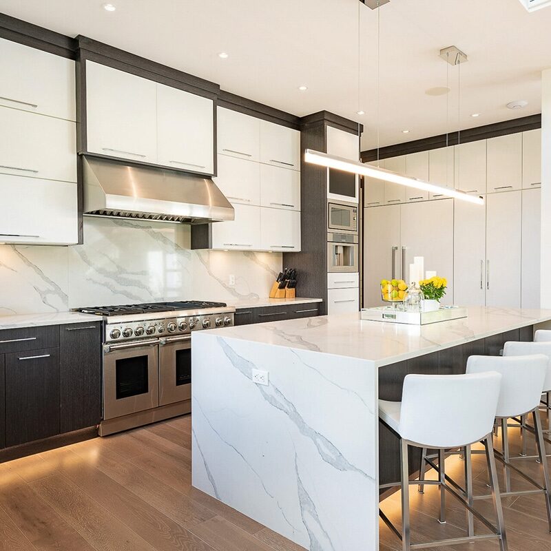 Modern kitchen with white and dark cabinets, marble countertops, a stylish stone kitchen backsplash, stainless steel appliances, a large island with four white barstools, and decorative yellow accents.