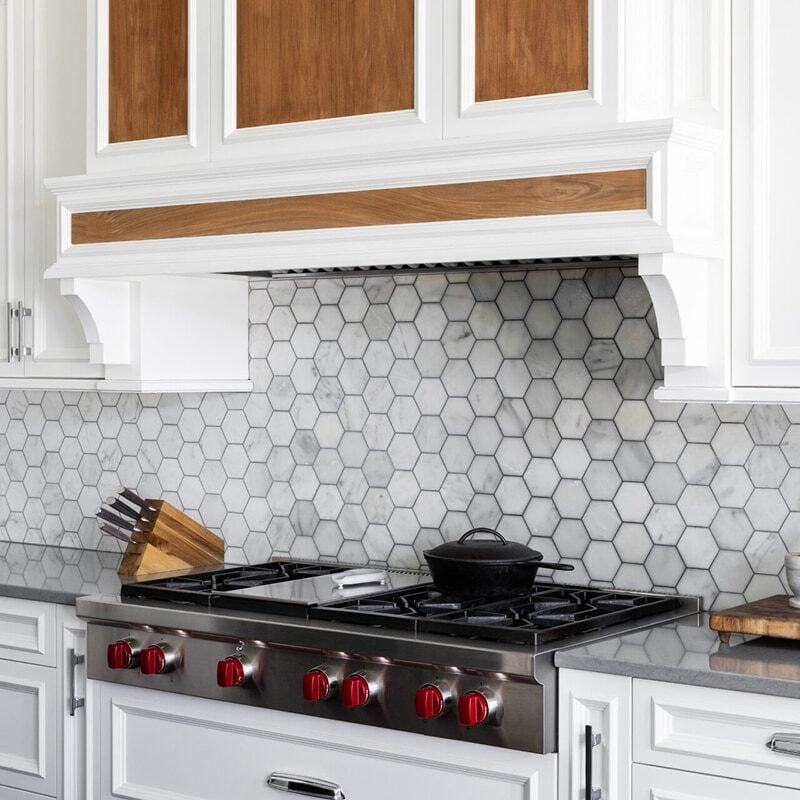 Modern kitchen with white cabinets, gray stone countertops, a hexagonal tile kitchen backsplash, stainless steel gas stove with red knobs, a black pot, knife block, and a small wooden cutting board with kitchen items.