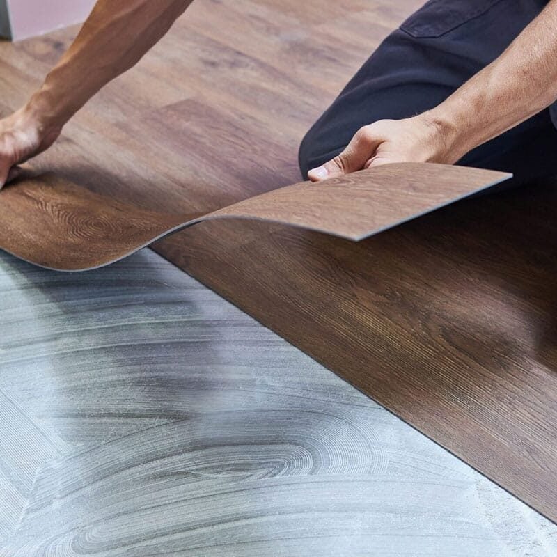 A person installs brown waterproof flooring, carefully placing a plank over adhesive on the subfloor, while wearing blue pants and kneeling on the ground.