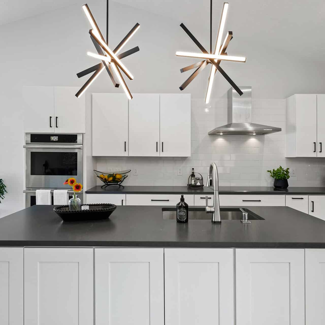 Modern kitchen remodel with white cabinets, black countertops, stainless steel appliances, and two geometric pendant lights above a large island with a sink. A window lets in natural light, and plants decorate the space.